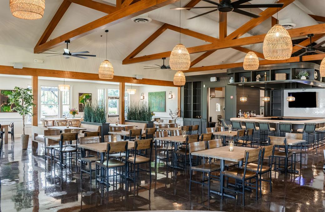 A restaurant with wooden beams on a tall ceiling, white walls, and wooden tables and chairs. Woven light fixtures hang from the ceiling.