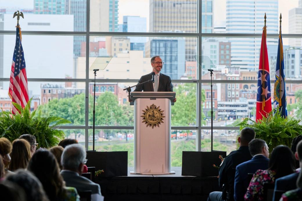 Nashville Mayor Freddie O'Connell in a suit and glasses smiles while speaking at a podium bearing the seal of Metropolitan Nashville-Davidson County, addressing a seated audience. The American flag and Tennessee state flag flank the stage, with ferns as décor. Floor-to-ceiling windows behind him reveal a downtown Nashville skyline and riverfront on a partly cloudy day.