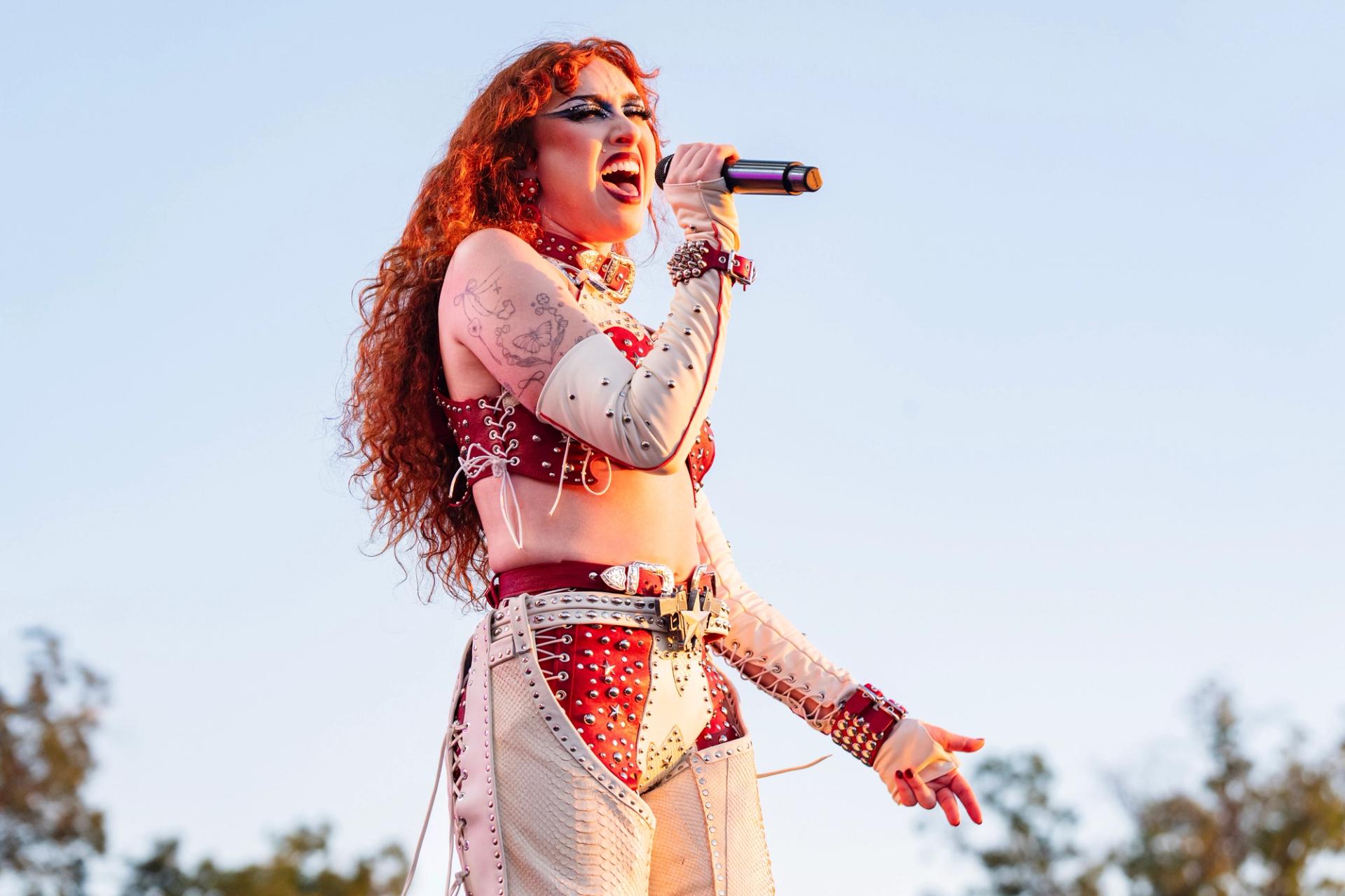 Chappell Roan wearing white and red leather chaps and a red leather top with studs. Her hair is bright red and she's singing into a microphone.