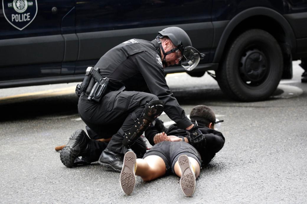 SPD officer with a gun on his hip and a helmet on kneels next to a person he is arresting who is laying on the pavement belly down with their hands pinned behind their back.