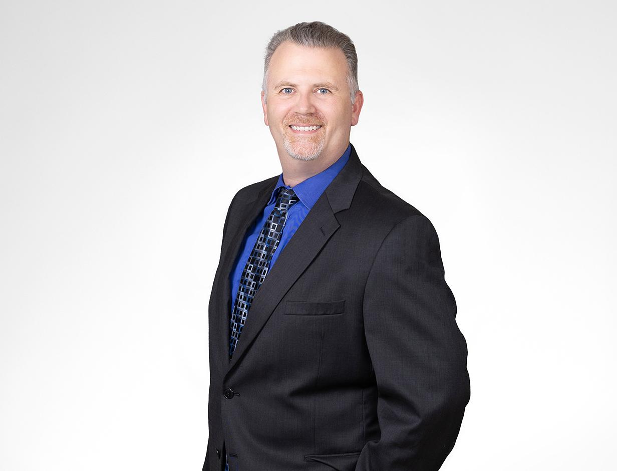 A photo of a smiling man wearing a darkly colored suit. He has on a bright blue button-down shirt underneath his blazer.