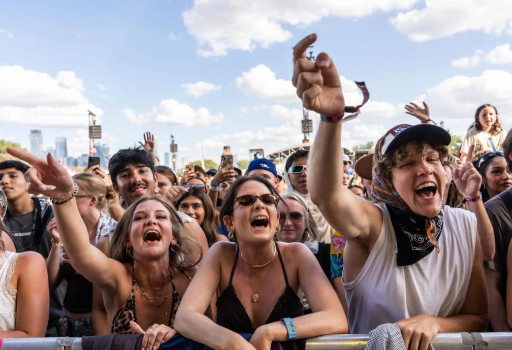 Concert attendees lined up at a barricade, screaming with their arms in the air. A blue sky is in the background.