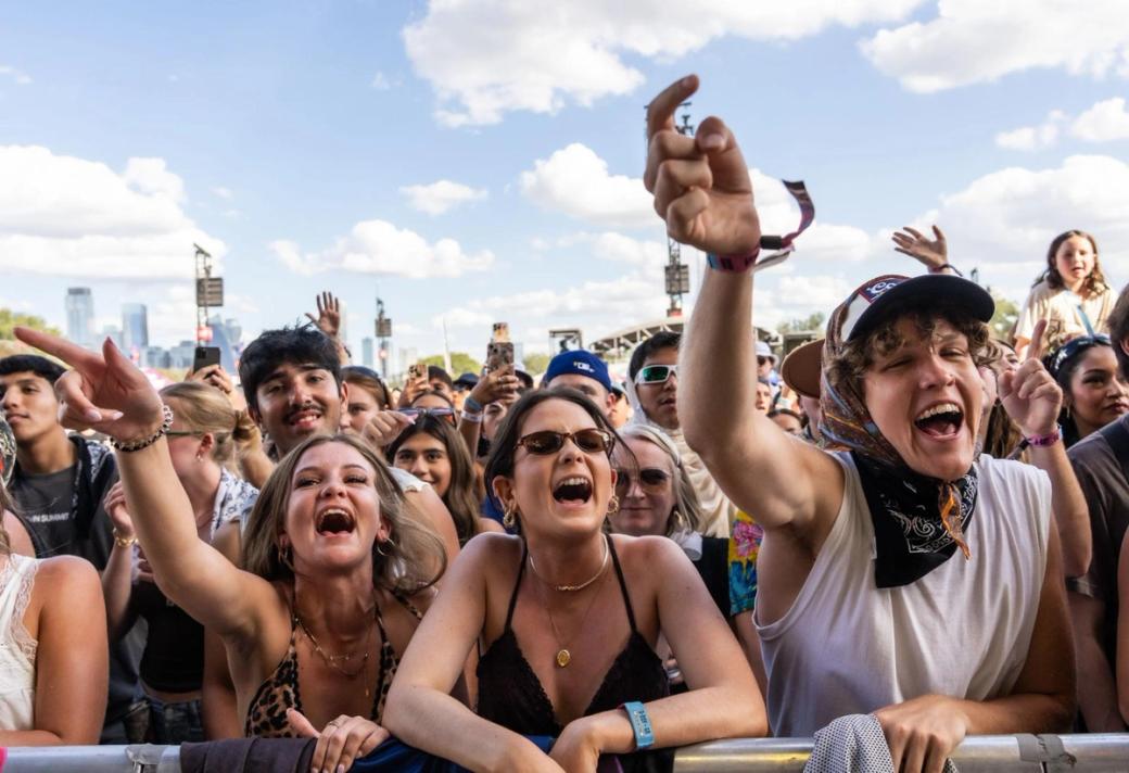 Concert attendees lined up at a barricade, screaming with their arms in the air. A blue sky is in the background.