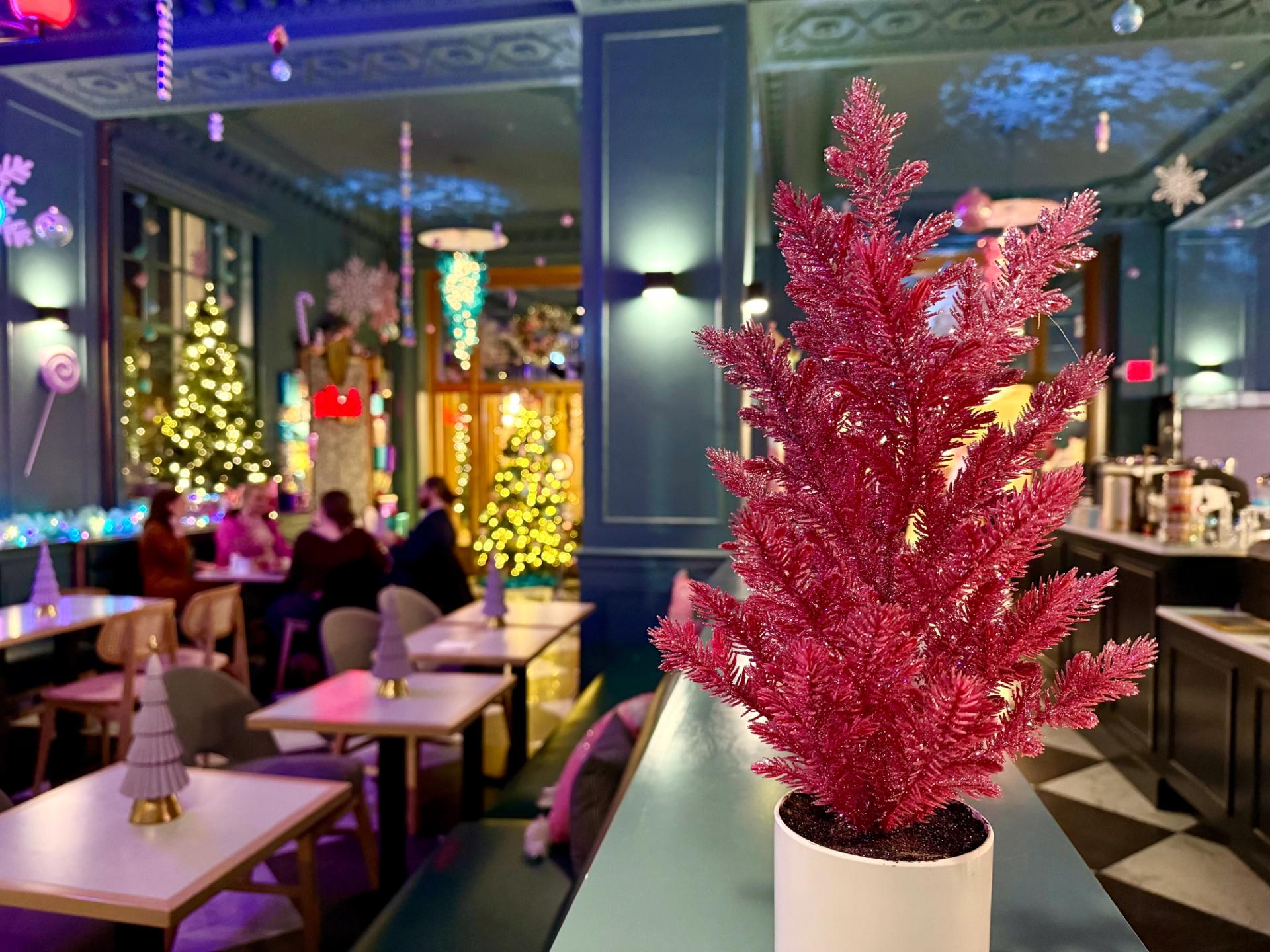 a pink plastic christmas tree in front of a pastel colored bar with people dining and christmas trees in the background