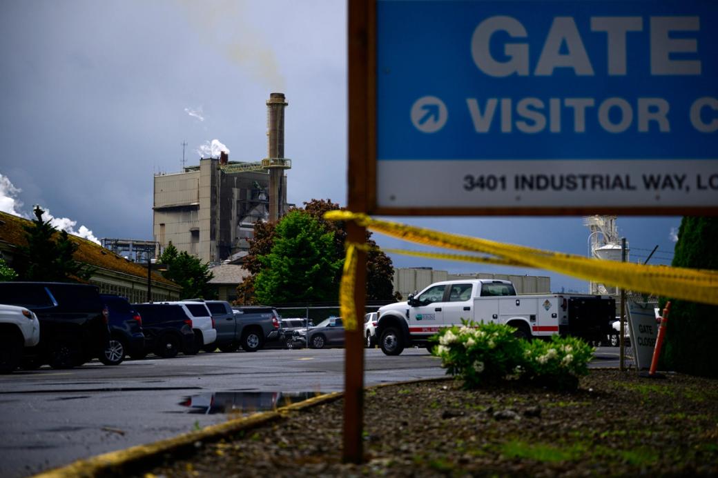 An industrial facility emitting smoke from a tall smokestack, viewed from a parking lot behind yellow caution tape. A partially visible blue sign reads 'Gate — Visitor — 3401 Industrial Way.