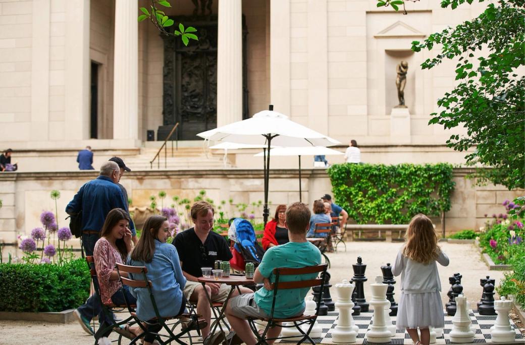 People sitting at a table outside the Rodin while a child plays with large chess pieces