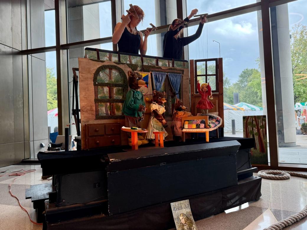 Two puppeteers control the strings for three bears and Goldilocks marionettes. They stand on a small black stage, with a set that looks like the inside of a house. They're performing in front of a wall of windows. Margaret Kingsbury Margaret Kingsbury 11:10 AM Today   Kyle, this day of events has been edited down.