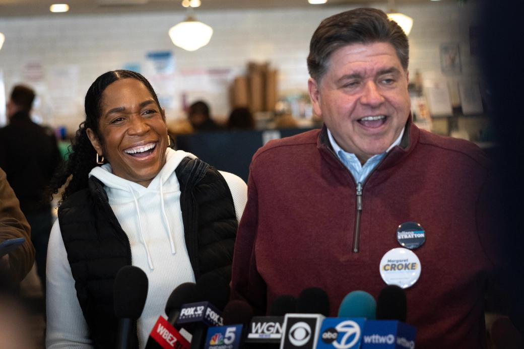 Man and woman smile in front of seven microphones outfitted with various news organization's branding on them