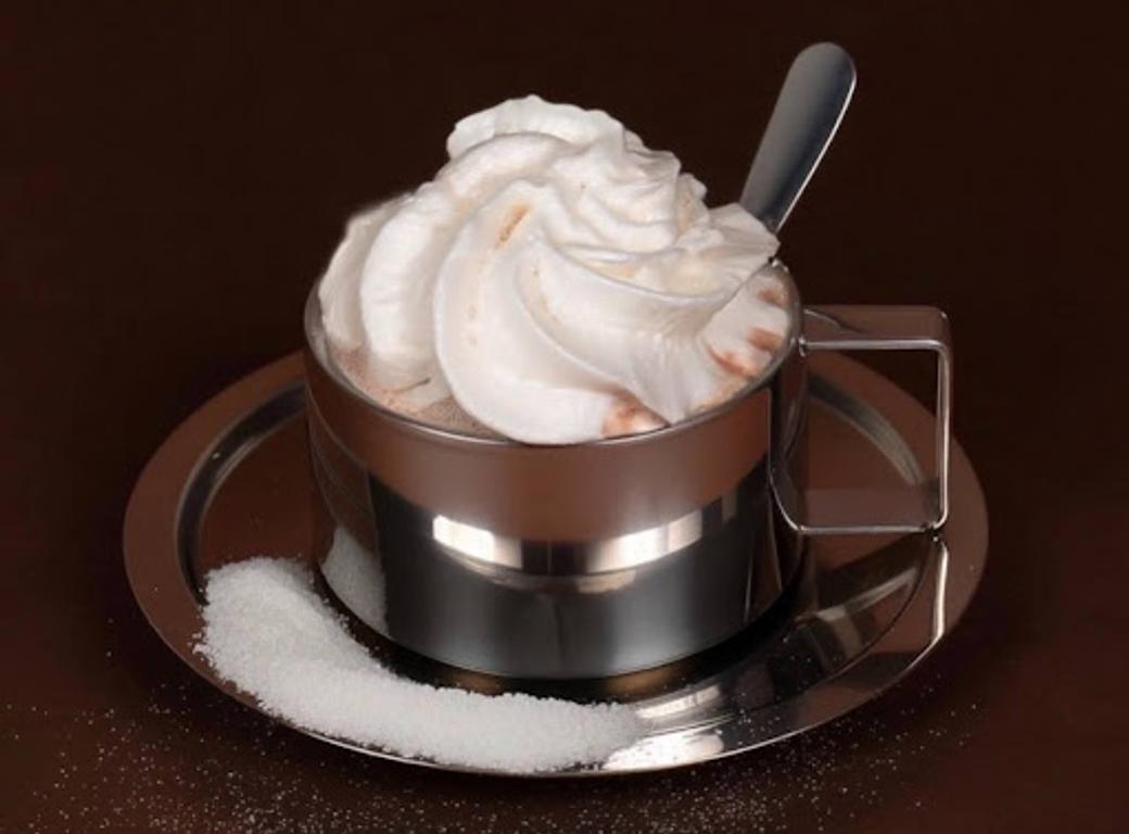 A chrome mug of hot chocolate with whipped cream, served on a chrome plate with a spoon and sprinkled sugar. 