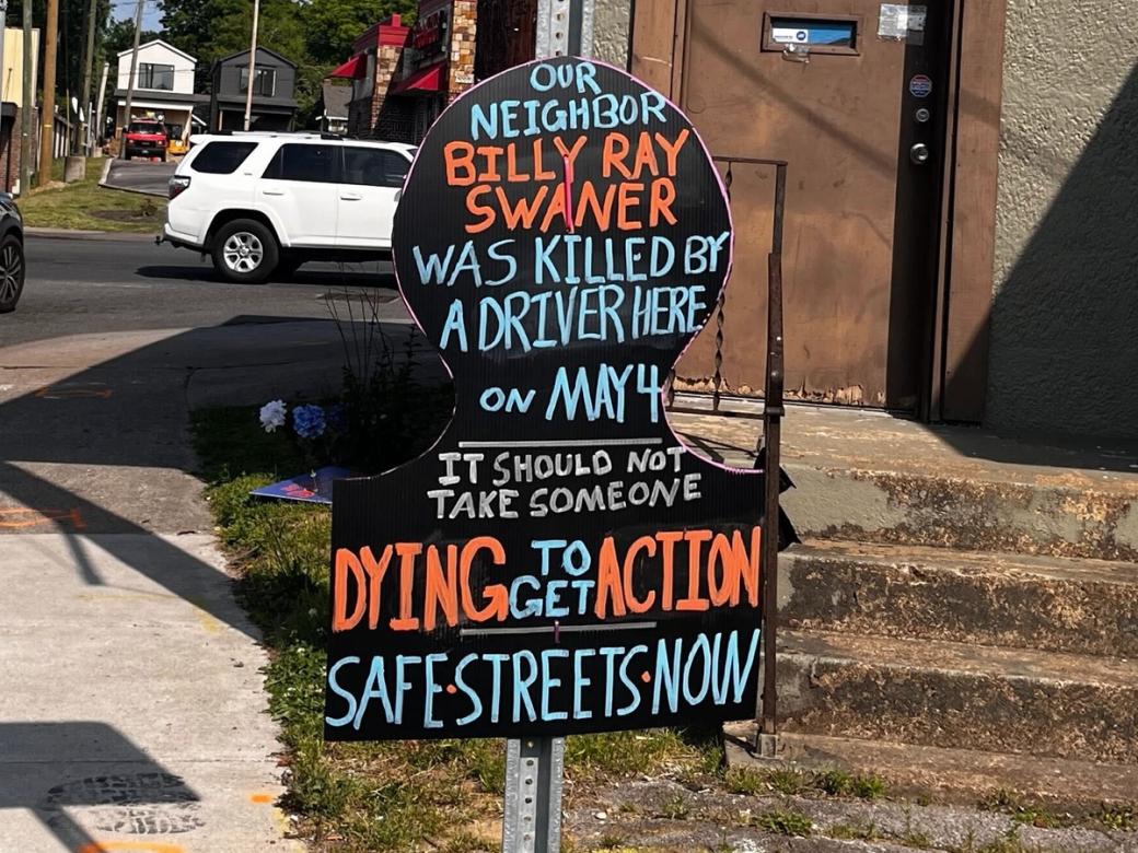 A black sign on a stop sign with blue, red, and gray wording. Text reads: Our neighbor Billy Ray Swaner was killed by a driver here on May 4. It should not take someone dying to get action. Safe streets now.