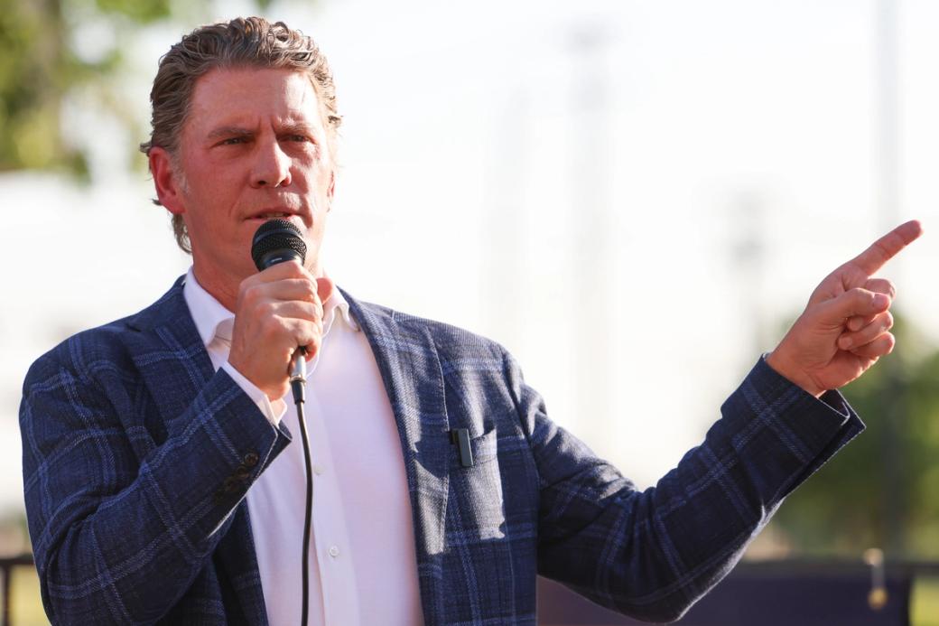 A man wearing a plaid suit jacket holds a microphone in one hand and points with the other hand.