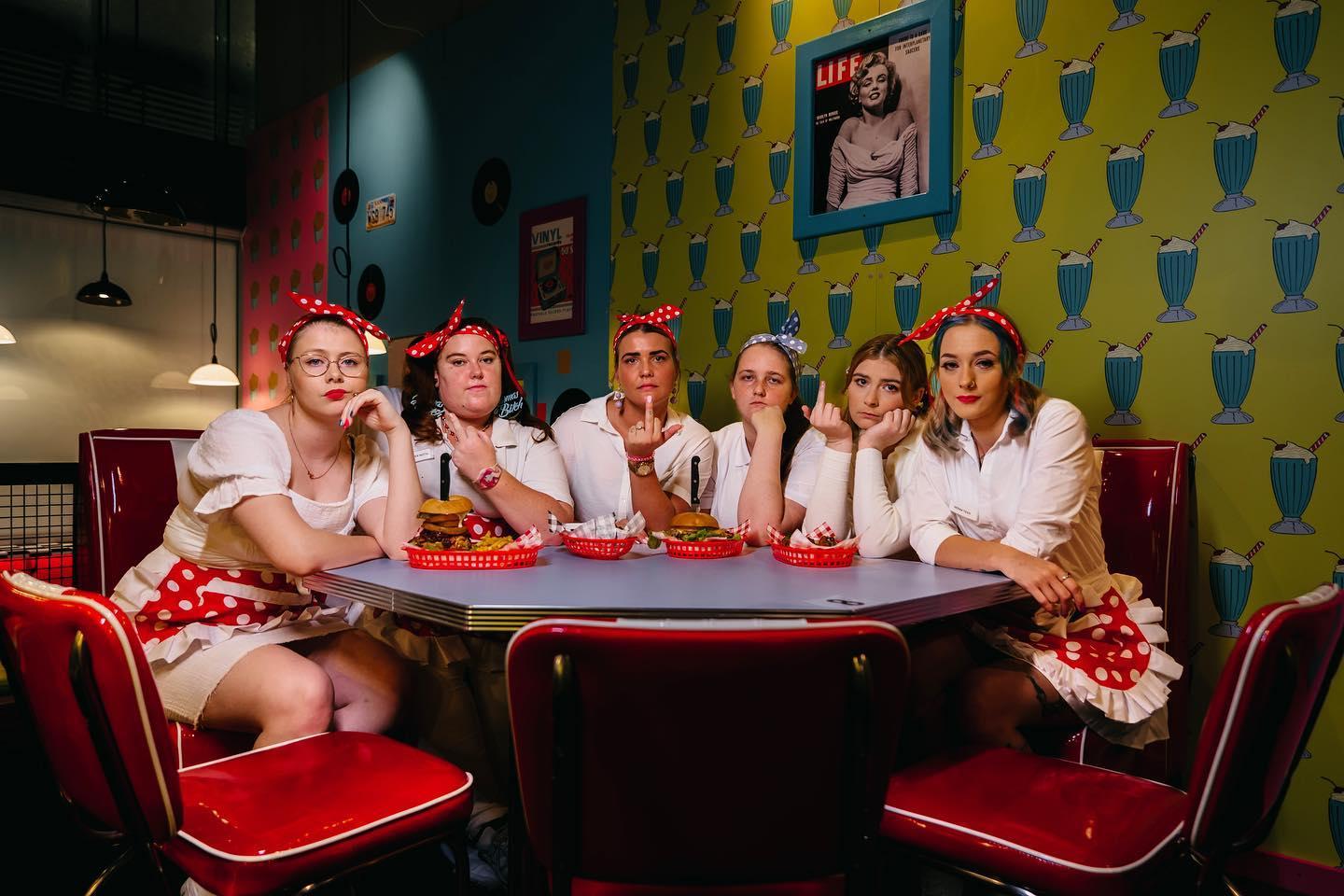 A group of angry-looking waitresses sit at a 1950s-style diner booth