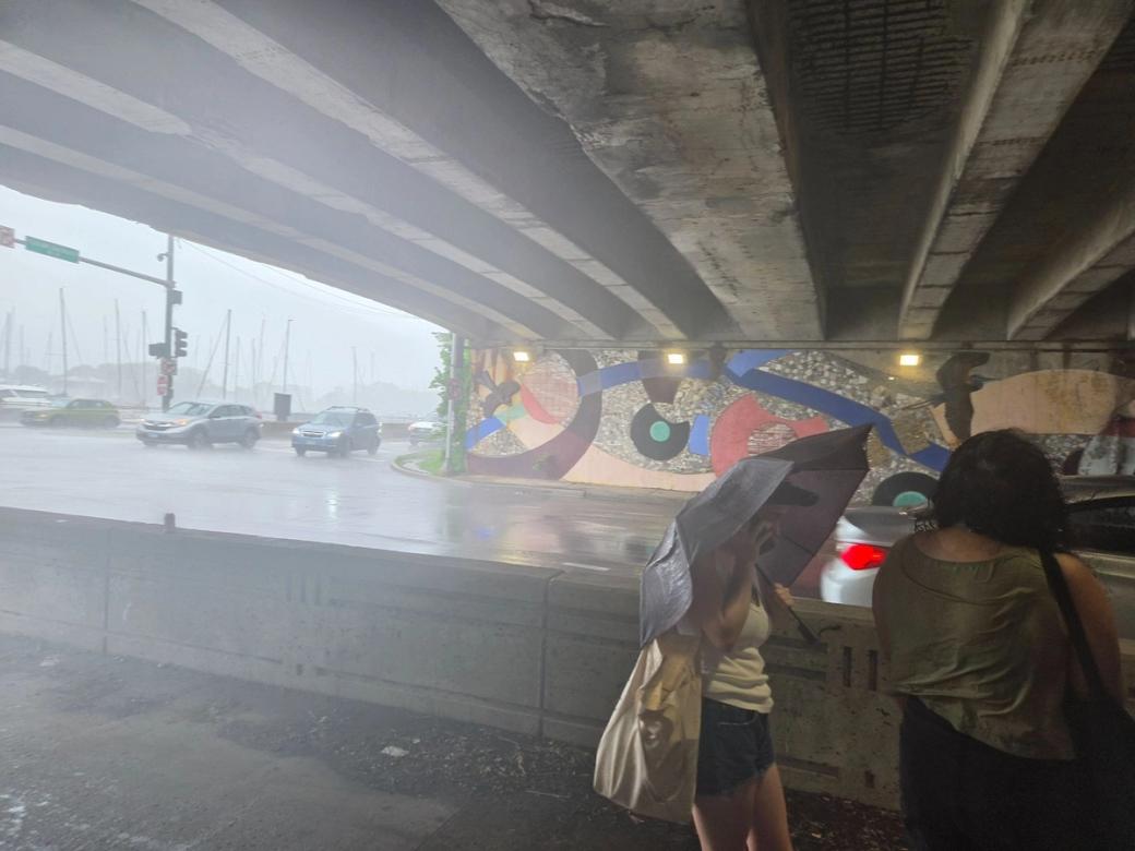 Two women under an overpass. One of which is holding an umbrella