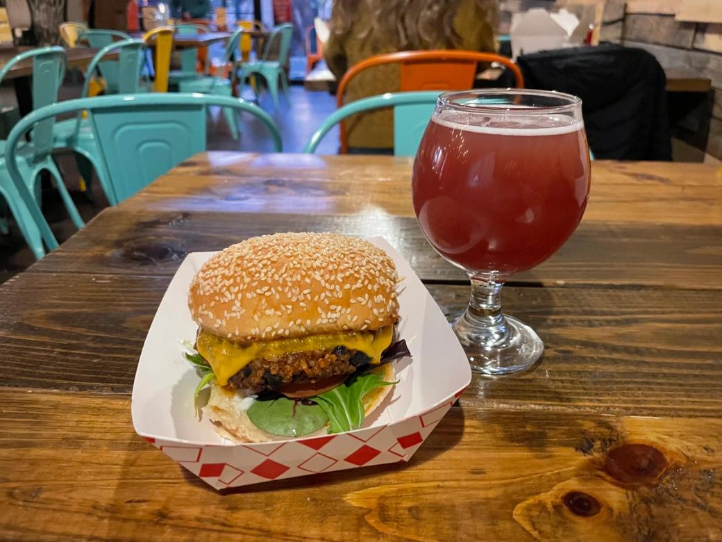 A red sour beer in a glass sitting next to a veggie burger on a sesame seed bud