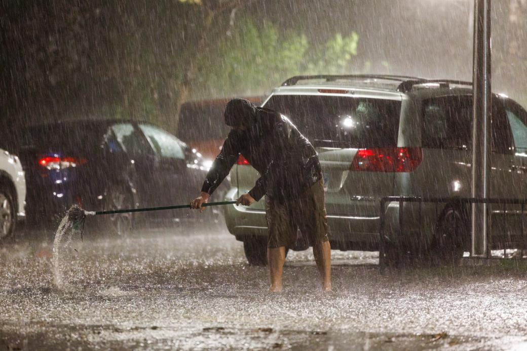 A man unclogging a drain in the road. The rain is pouring heavy and the road looks flooded