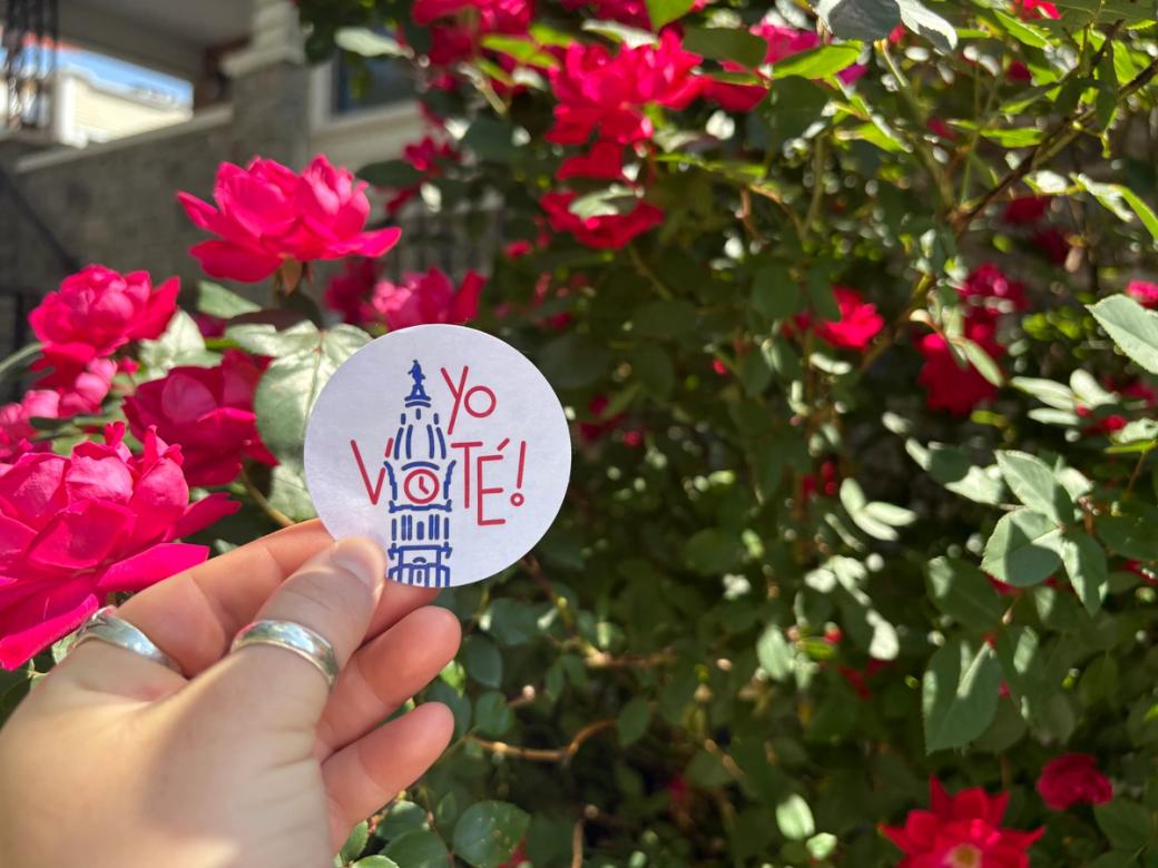 A hand holds a "Yo Voté!" sticker with an illustration of Philadelphia's City Hall in front of a bush of flowers