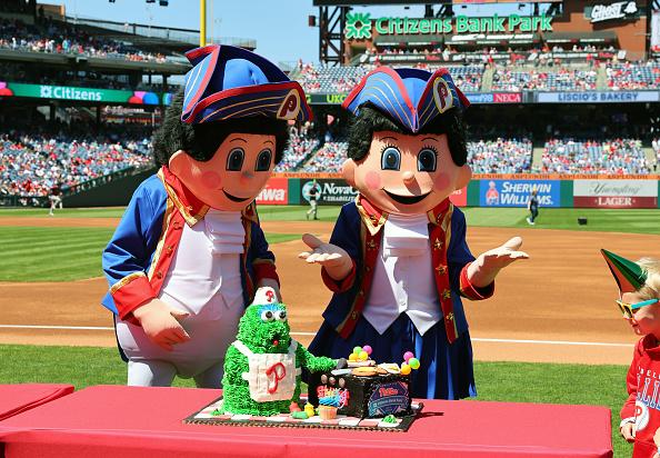 Two human-like caricatures of people in colonial clothes stand on the Phillies home baseball field with a cake in front of them of the current Phils mascot, the Phanatic.