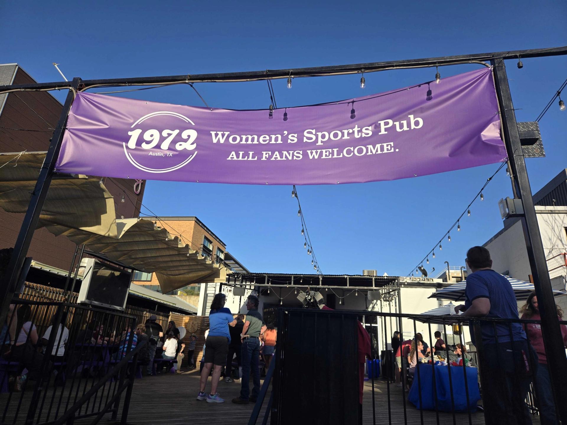A purple banner hanging over an outdoor patio at a bar that reads: "1972 women's sports pub. All fans welcome."