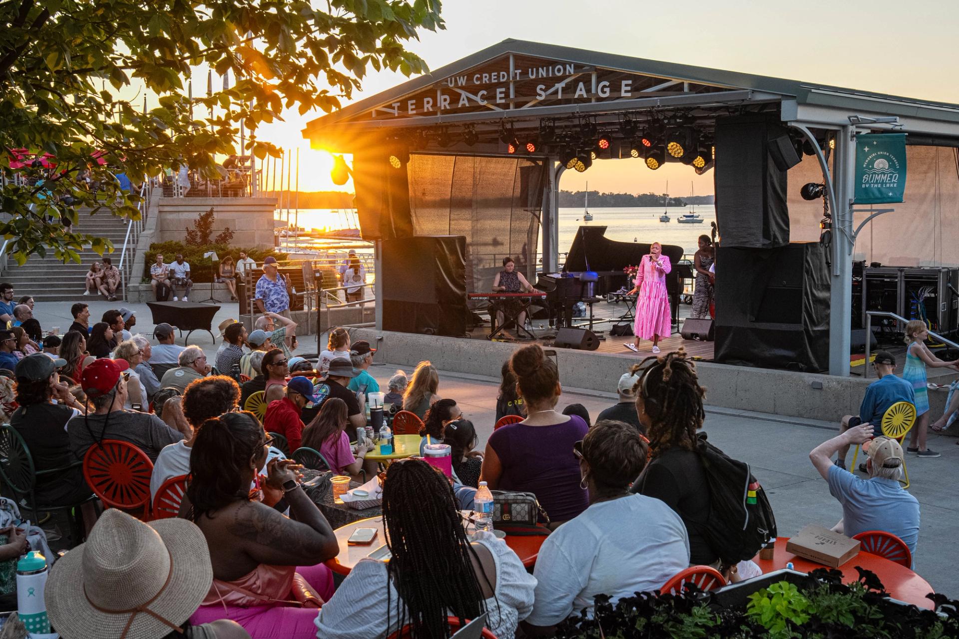 A woman in a pink outfit sings on an outdoor stage before a crowd.