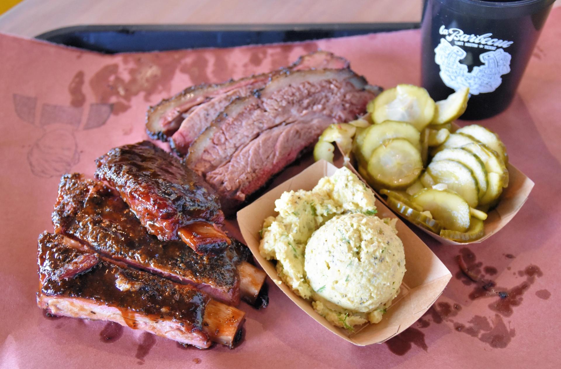 A plate of a barbecue that includes ribs and brisket. A brown basket of pickles and a brown basket of potato salad are also on the plate. 