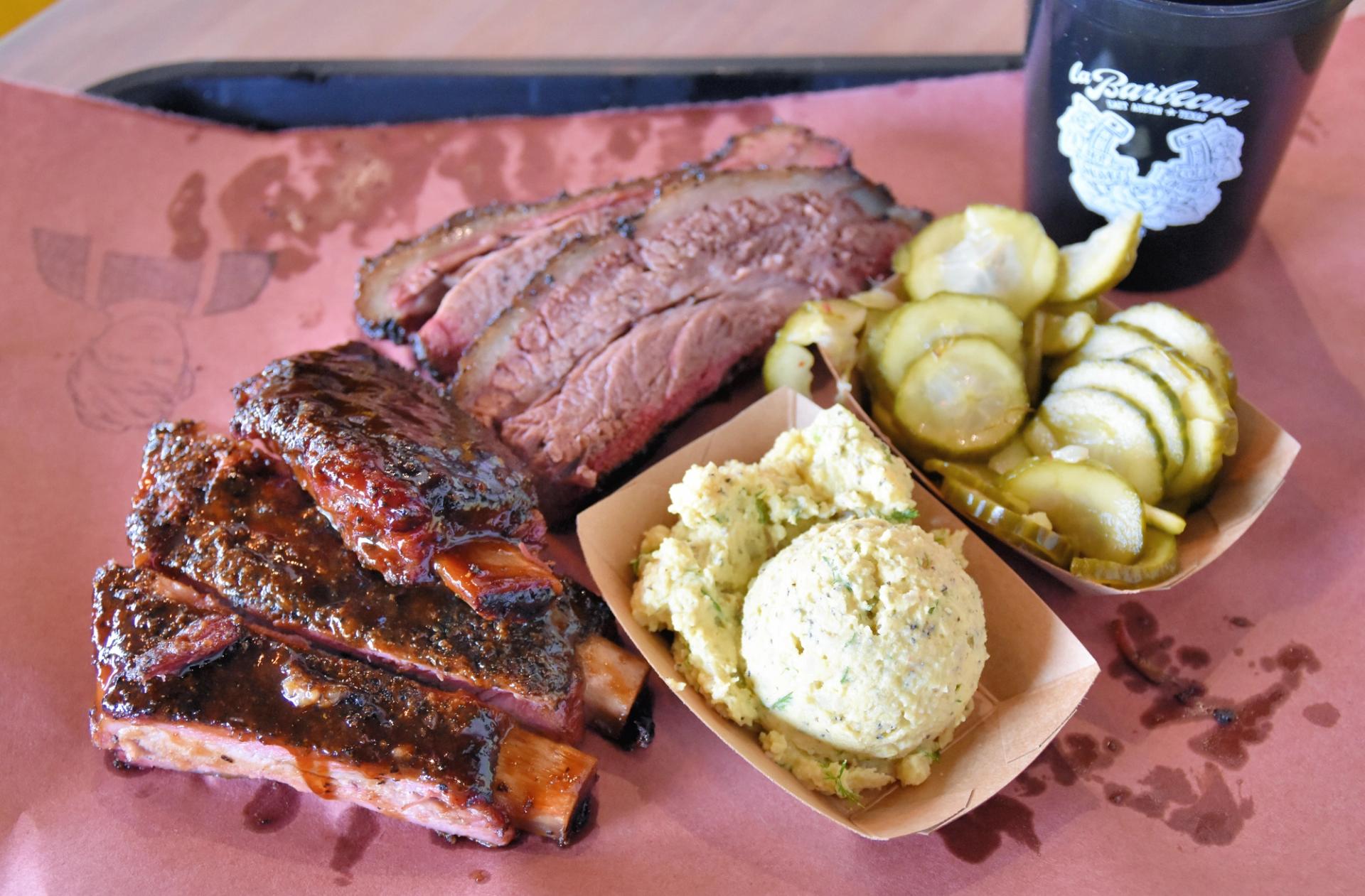 A plate of a barbecue that includes ribs and brisket. A brown basket of pickles and a brown basket of potato salad are also on the plate.