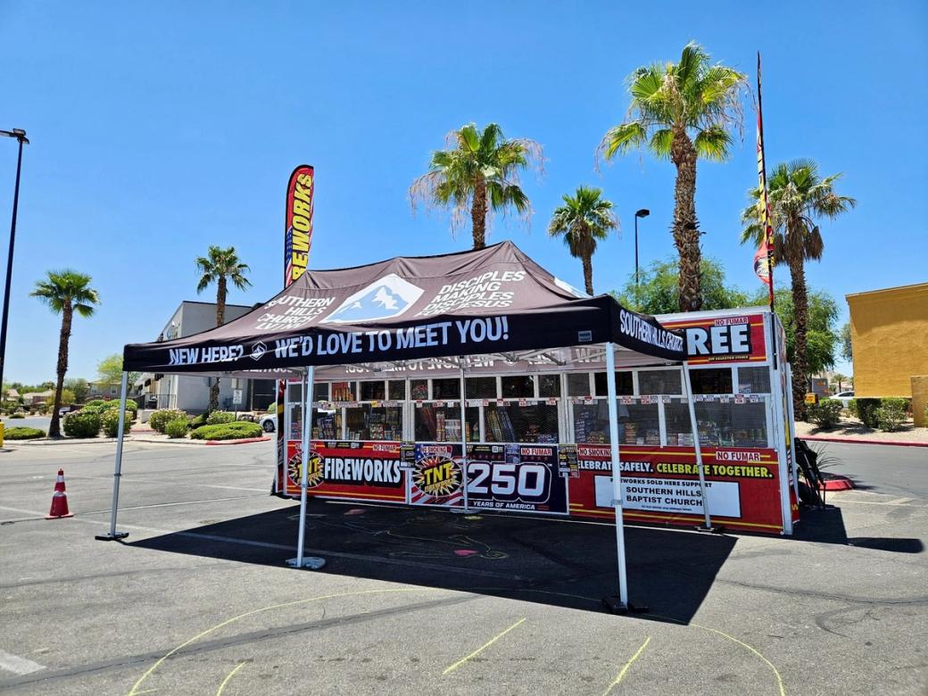 A fireworks stand with "FIREWORKS" and "250" written on it. In front is a brown awning reading "NEW HERE? WE'D LOVE TO MEET YOU"