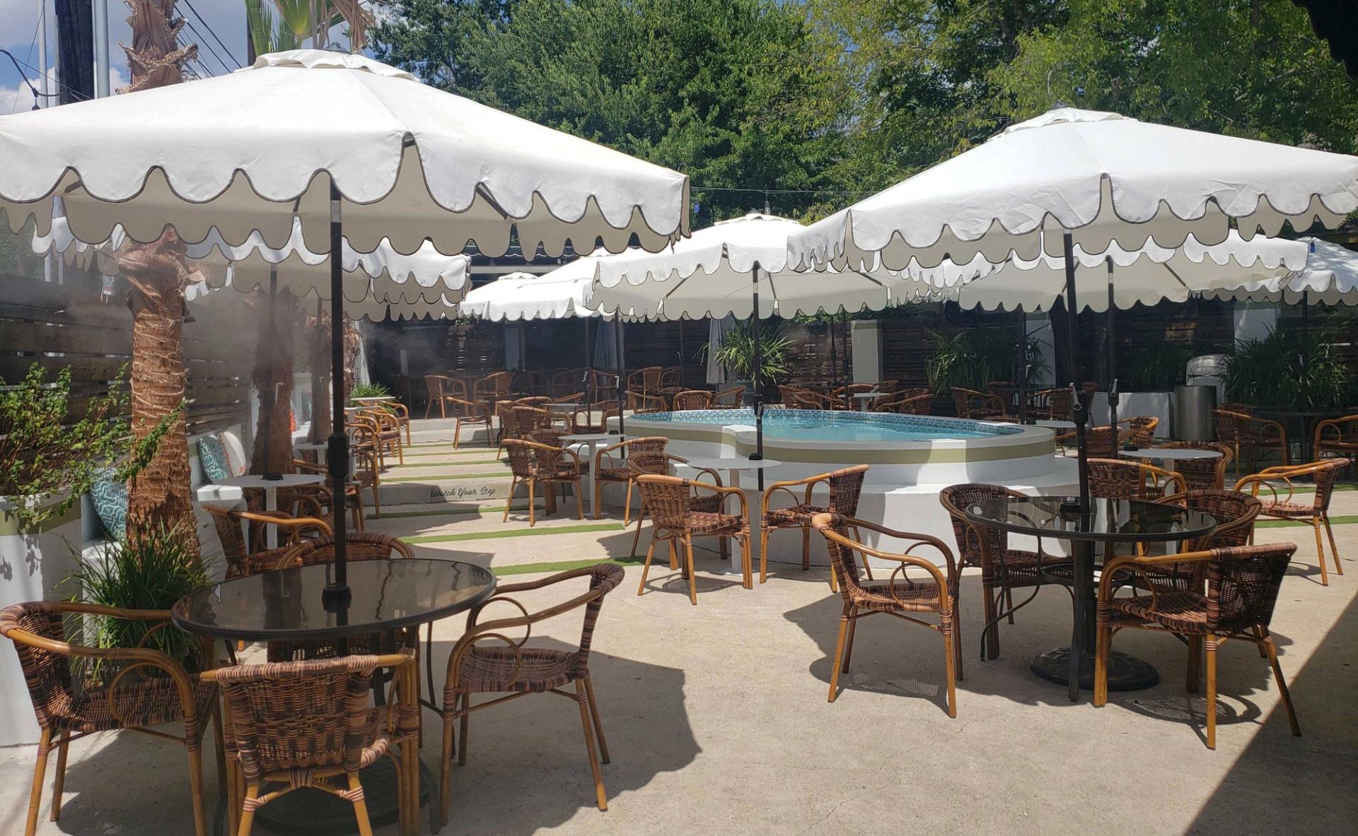 Patio tables with umbrellas and chair populate a cement, outdoor space. A small pool sits in between the tables.