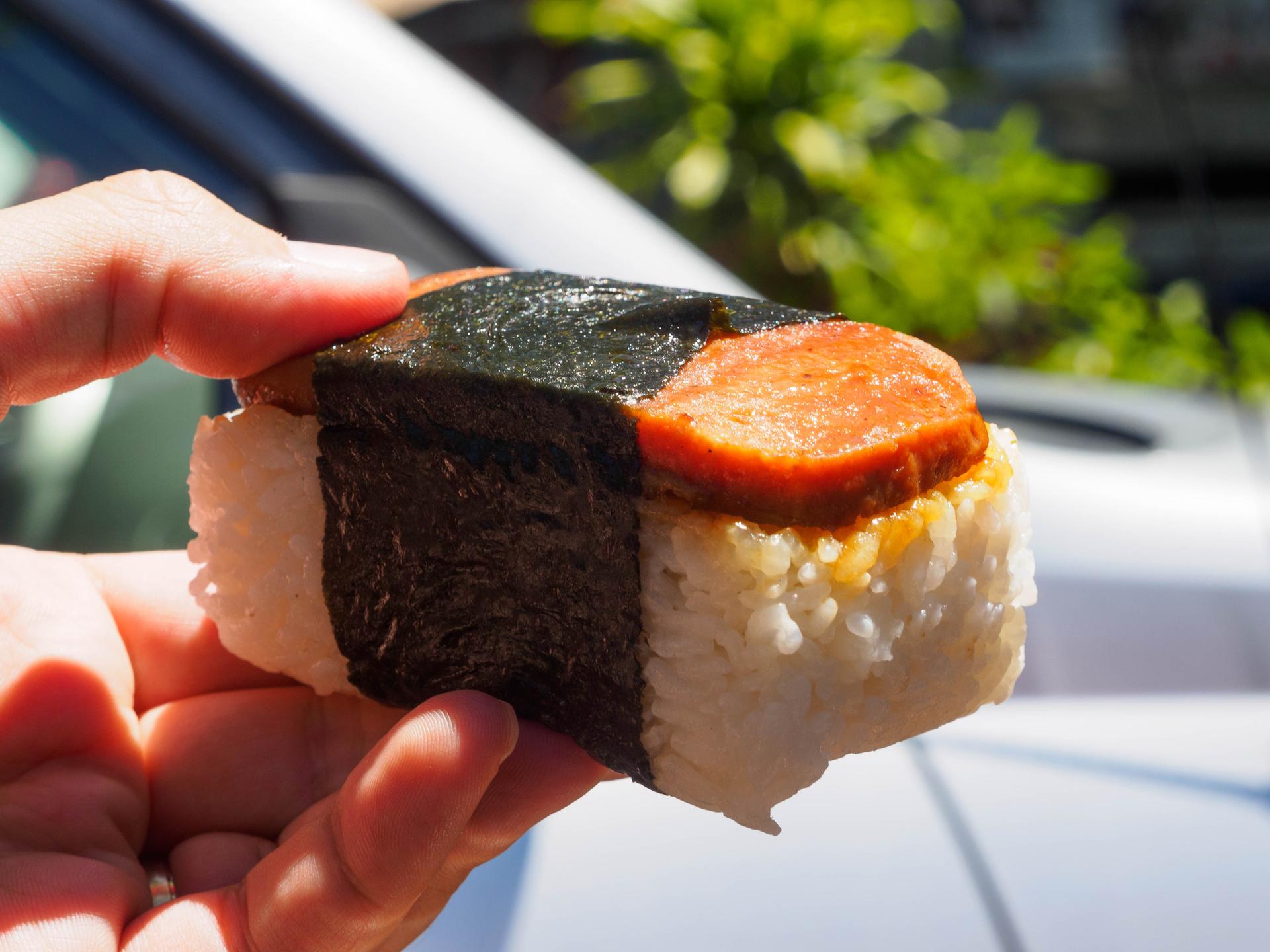 A pale hand holds a piece of spam musubi — a roughly 3 inch block of rice with a slice of seared spam on top, wrapped around the center with a ribbon of dried seaweed.