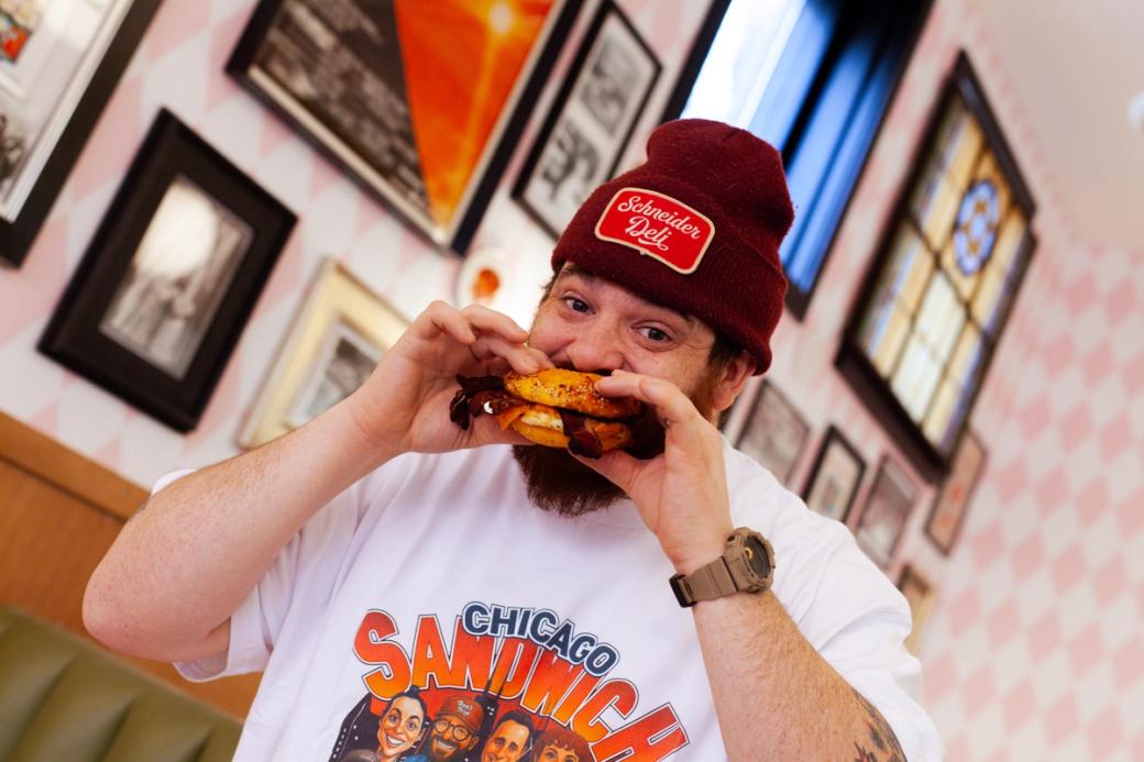 Bearded man wearing beanie and t-shirt holds sandwich up to his mouth indoors