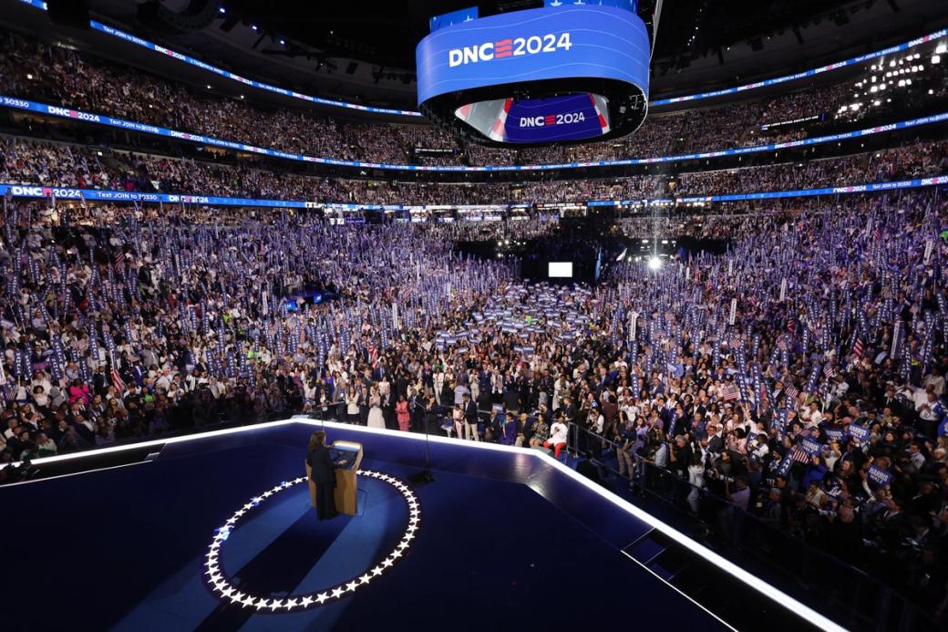 A person standing at a podium addressing a massive crowd of people. There' a sign on the jumbotron that says "DNC 2024"