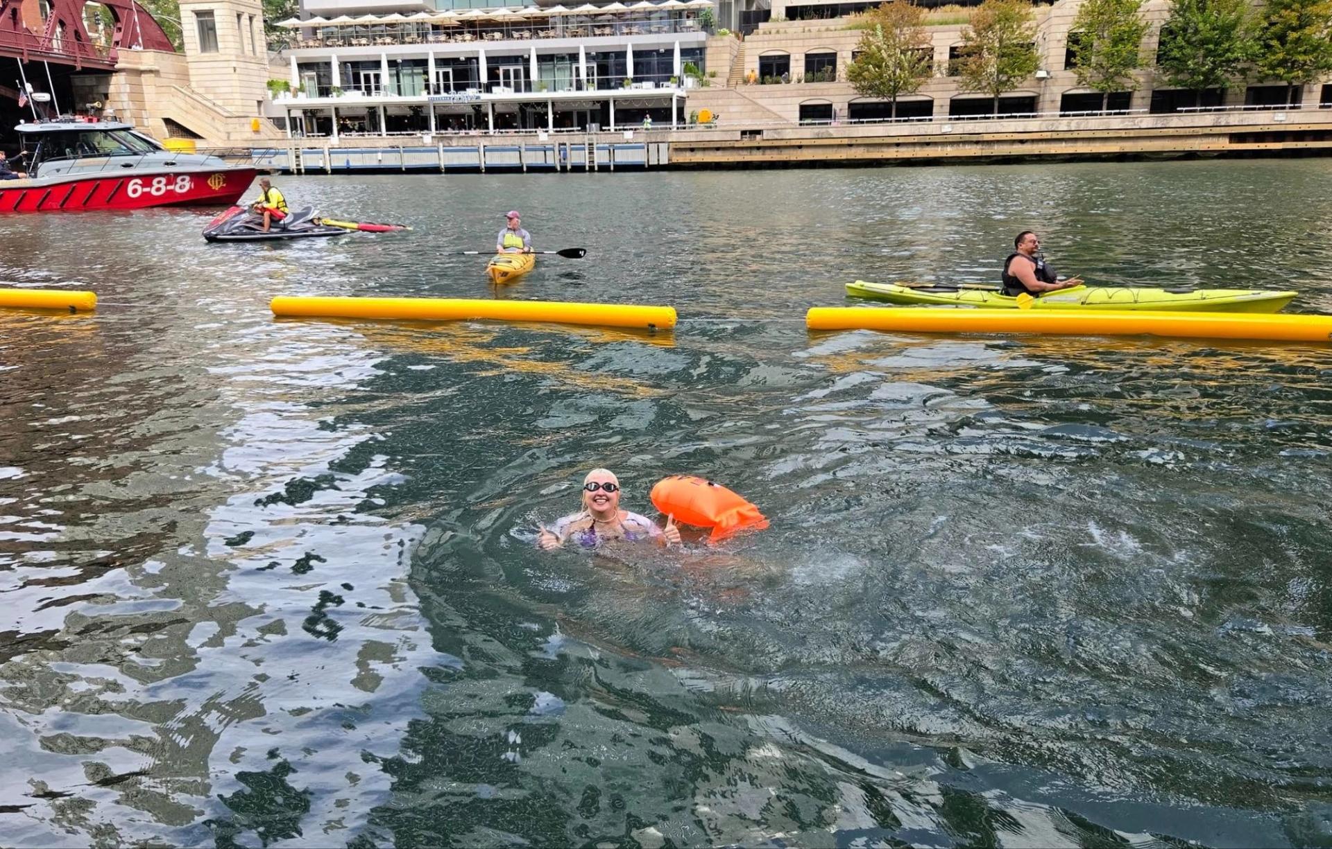 A woman wearing goggles and holding a flotation device in a river. There are canoes and a boat in the background