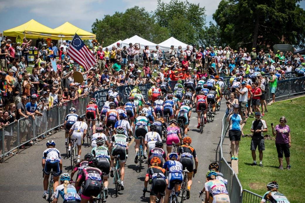 A large pack of competitive cyclists climbs the Lemon Hill course at the 2015 Philadelphia Cycling Classic, cheered on by crowds lining both sides of the barricaded road.