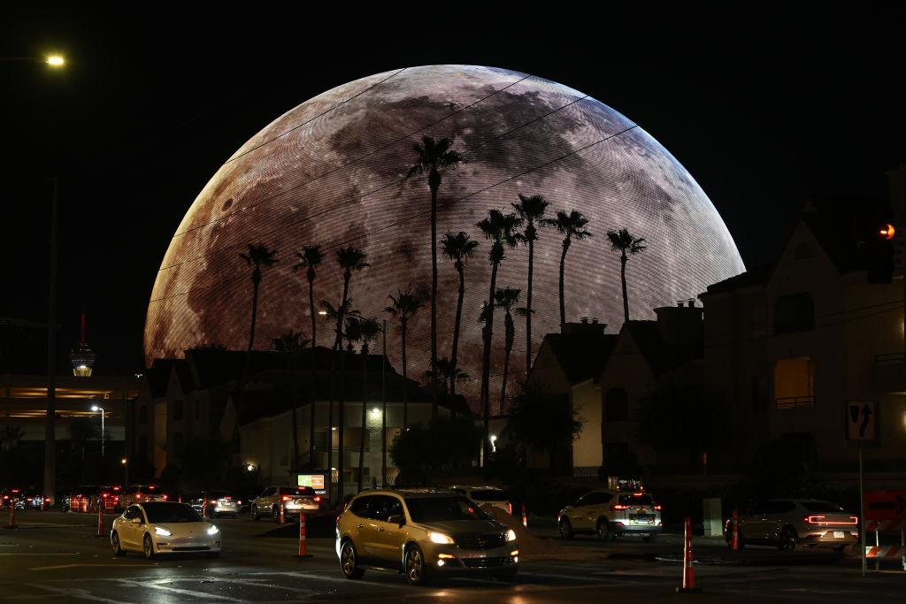 An evening street view of the Sphere in Las Vegas lit up as the moon. Traffic meanders in front of it.
