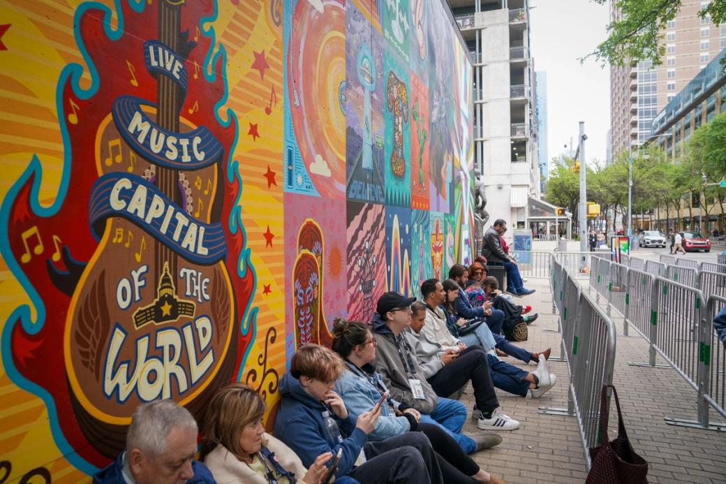 People sitting in a line along a wall with a colorful mural.