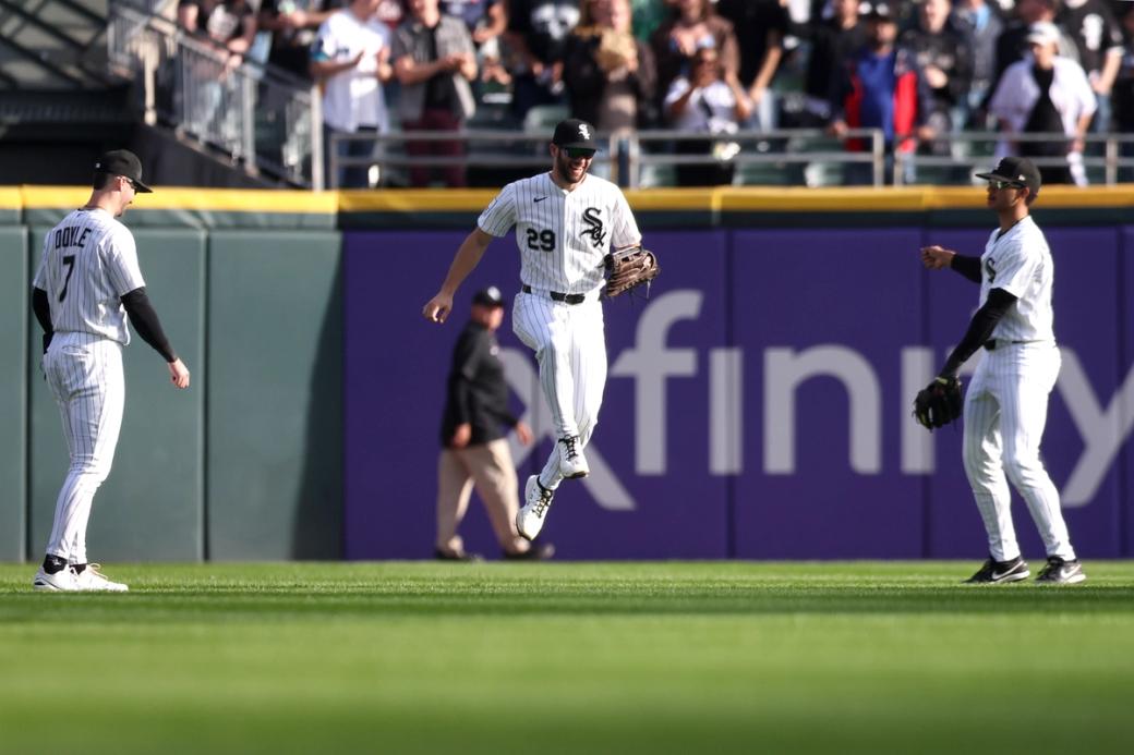 Three Chicago White Sox baseball players in white uniforms on the field during a game, with one player jumping toward two teammates near the outfield wall and a crowd in the stands behind them.