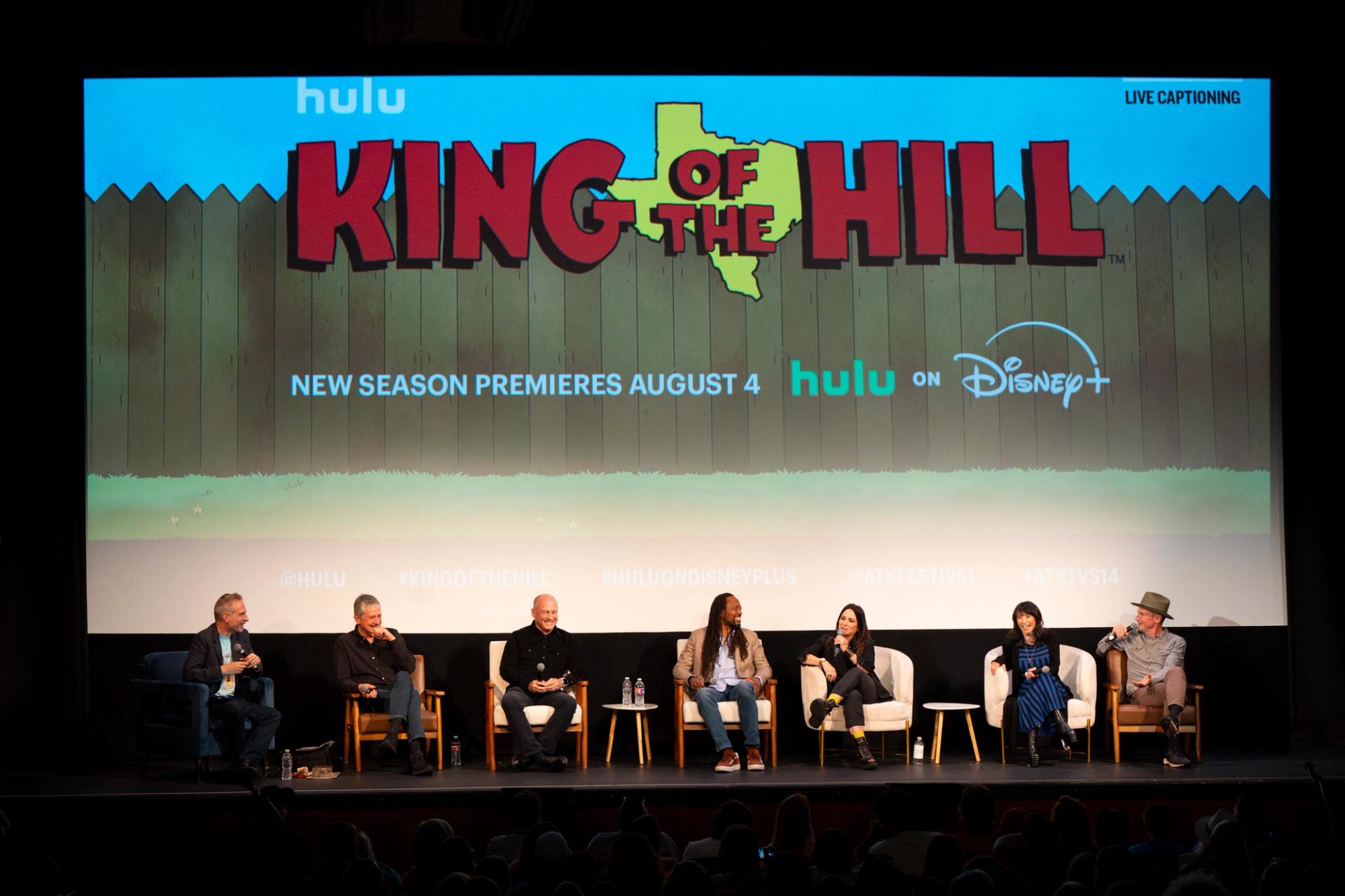 Seven people sitting in chairs on a stage with a "King of the Hill" sign above them.