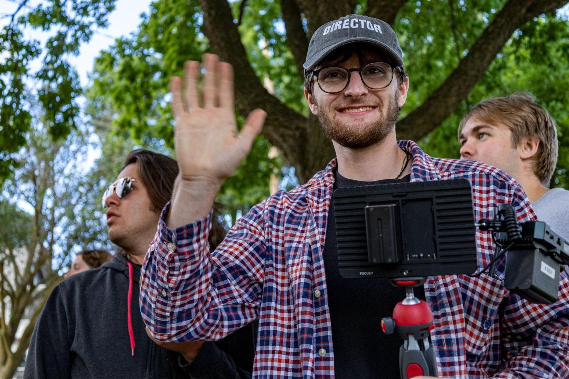 Filmmaker Corey Holl, wearing a hat that says “Director” smiles and waves to the camera as he checks a monitor.