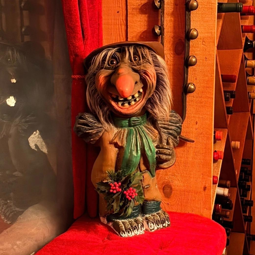 A grinning troll doll stands in front of  a wine shelf. 