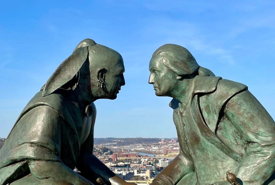 two sculpted bronze figures face each other in the foreground with a river, several bridges, and buildings in the distance behind them