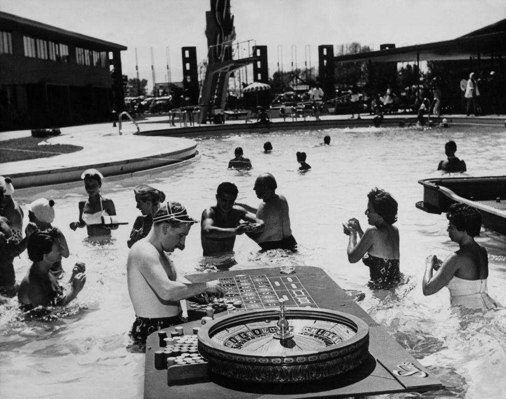 Black and white photo of people in a swimming pool, a roulette table in the forefront.