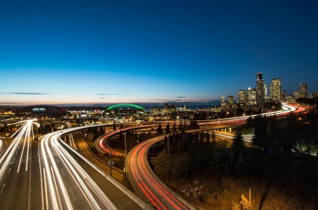 A long exposure photo of traffic across Seattle highways creates light streaks in white and red of car headlights and taillights with the city skyline in the background and a gradient dusky sky.