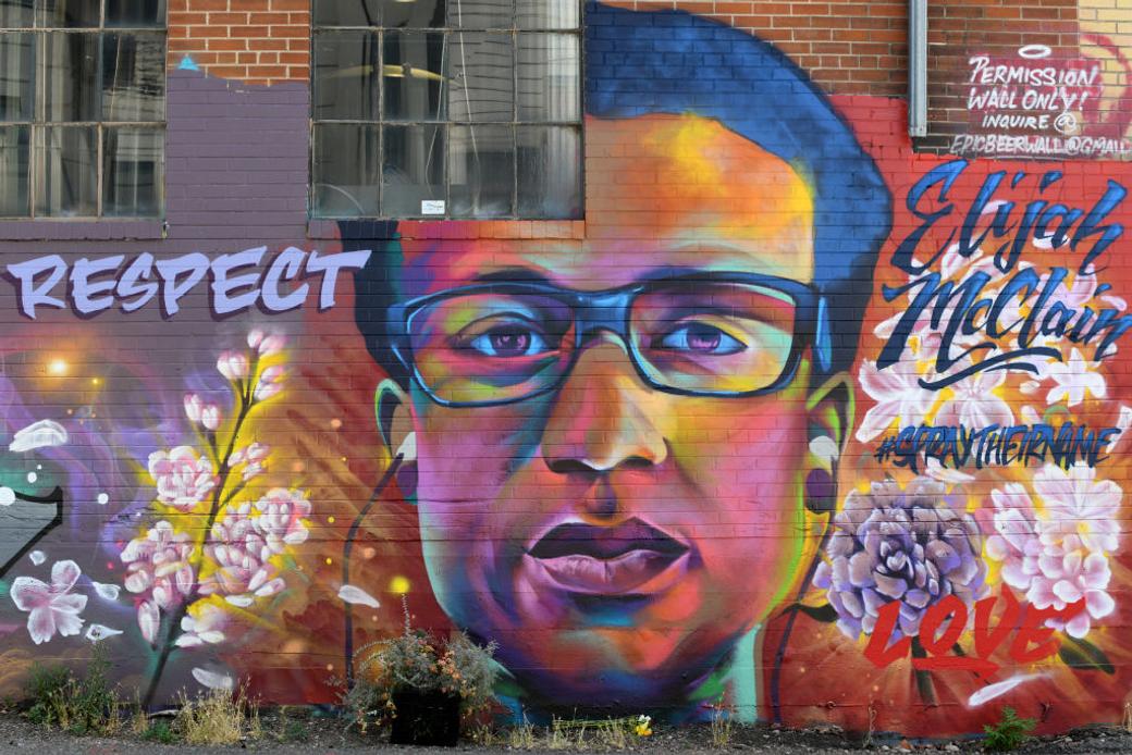 A vibrant mural on a brick building depicting a colorful portrait of Elijah McClain wearing glasses, surrounded by cherry blossoms. Text on the mural reads 'RESPECT,' 'Elijah McClain,' '#SayTheirName,' and 'Love.'