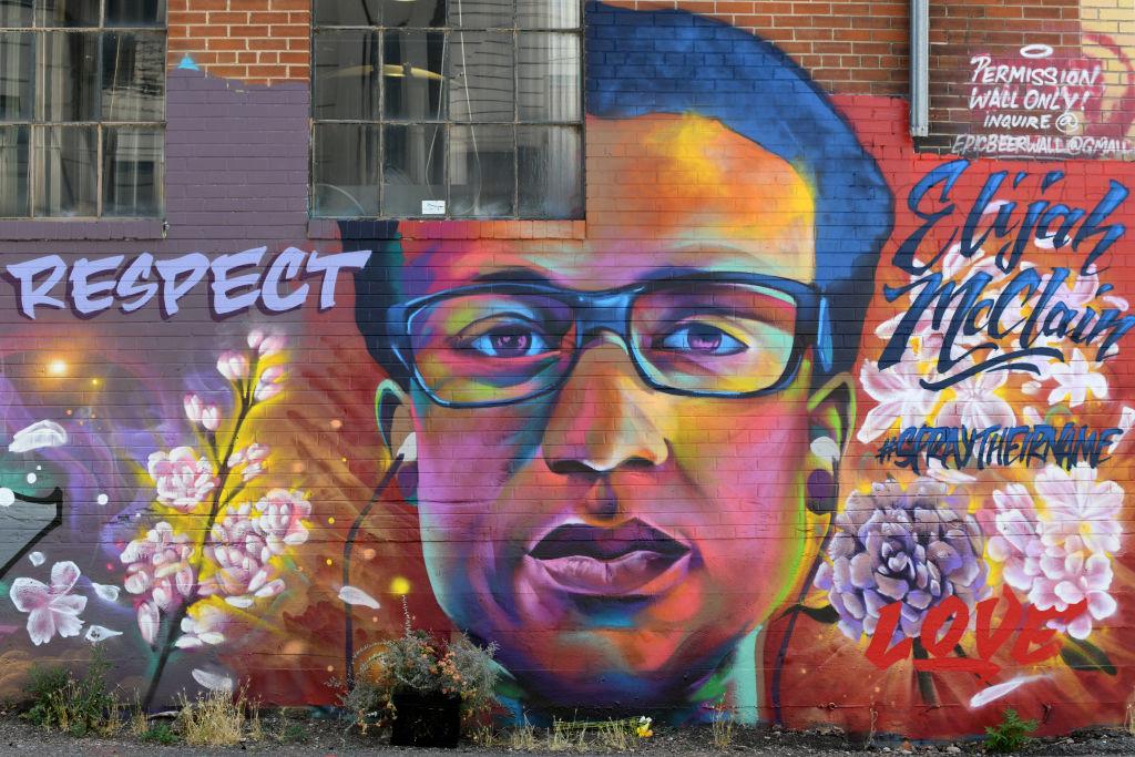 A vibrant mural on a brick building depicting a colorful portrait of Elijah McClain wearing glasses, surrounded by cherry blossoms. Text on the mural reads 'RESPECT,' 'Elijah McClain,' '#SayTheirName,' and 'Love.'