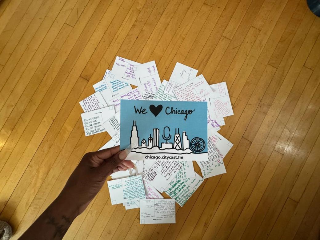 A hand holding a postcard that says "We love Chicago" There's an assortment of postcards under that postcard that are arranged in the shape of a heart