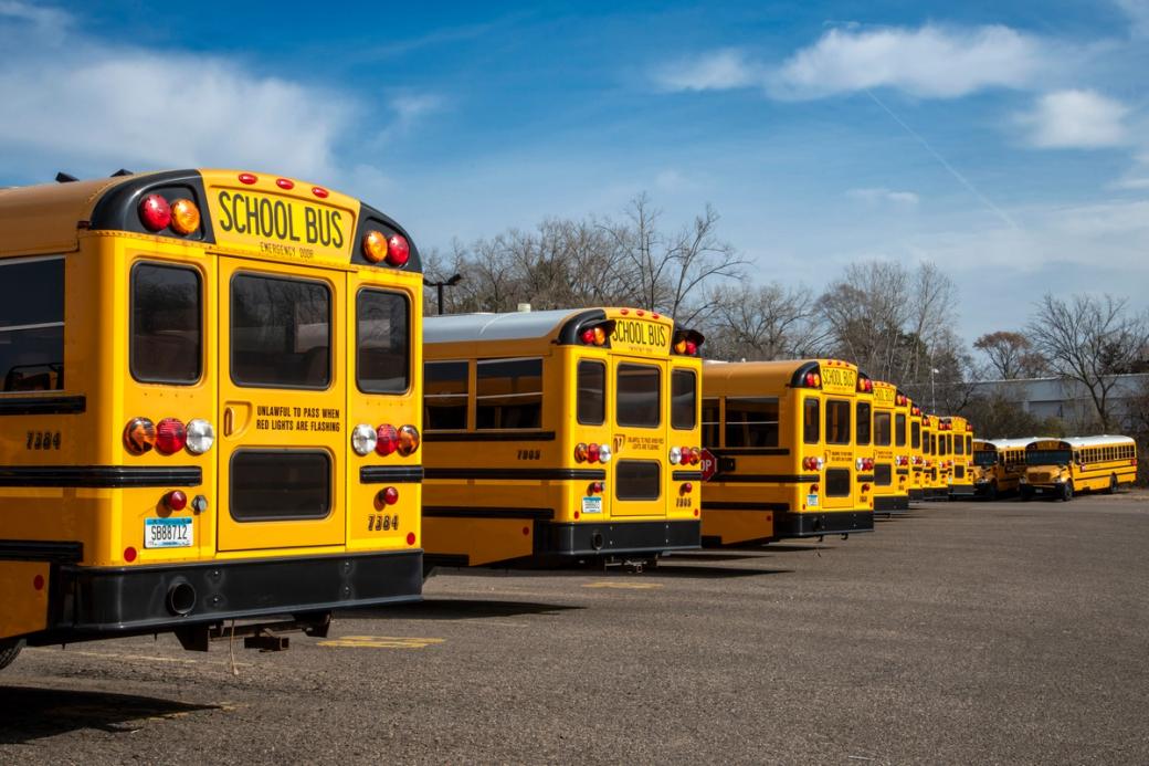 A row of parked yellow school buses, seen from behind, lined up diagonally in a paved lot under a blue sky with scattered clouds.