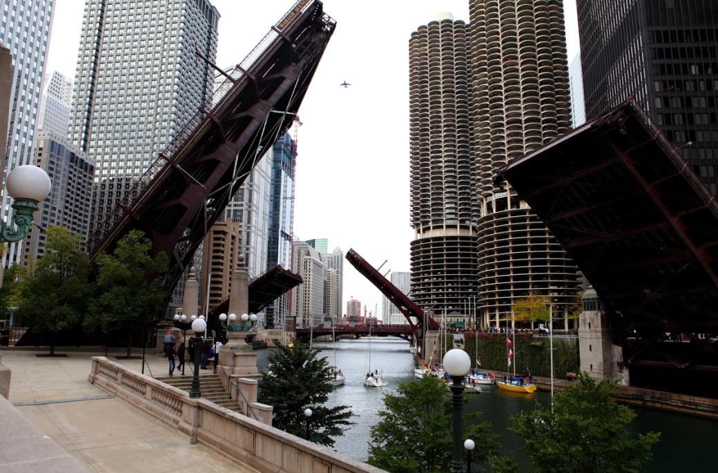 A view of the Chicago River with several raised drawbridges, surrounded by downtown skyscrapers, including Marina City’s distinctive cylindrical towers on the right. A pedestrian walkway and decorative street lamps line the river in the foreground.