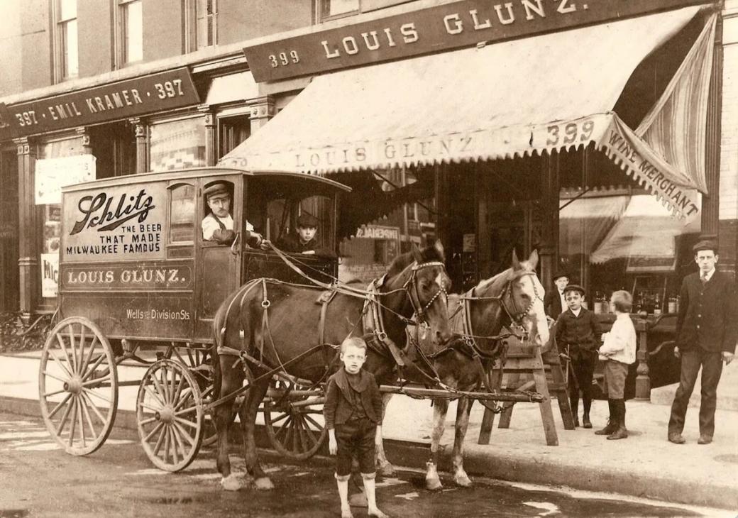 An old fading black and white photo of a boy standing in front of a man in a horse drawn carriage. On the side it says "Schlitz delivery cart"