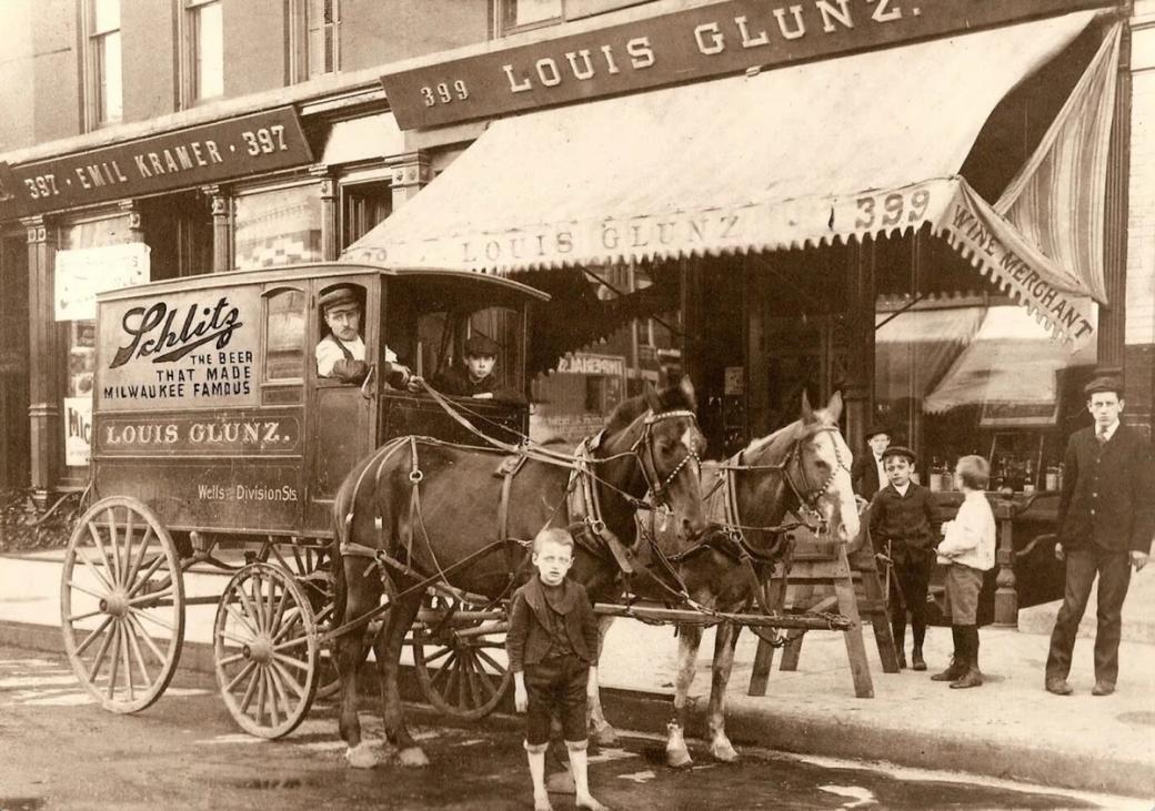 An old fading black and white photo of a boy standing in front of a man in a horse drawn carriage. On the side it says "Schlitz delivery cart"