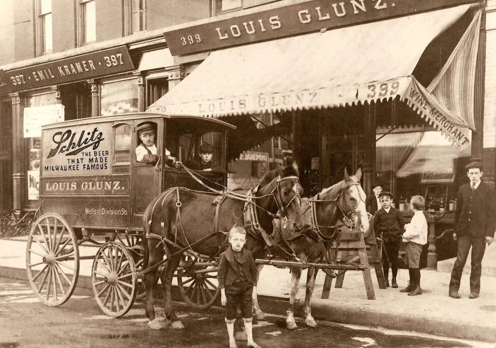 An old fading black and white photo of a boy standing in front of a man in a horse drawn carriage. On the side it says "Schlitz delivery cart"