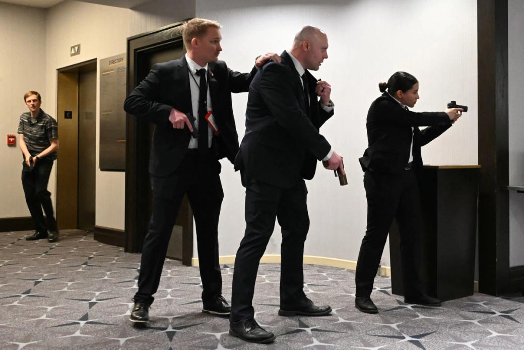 Three people in suits peer around corner with guns drawn, a fourth man stands in background with gun in hands